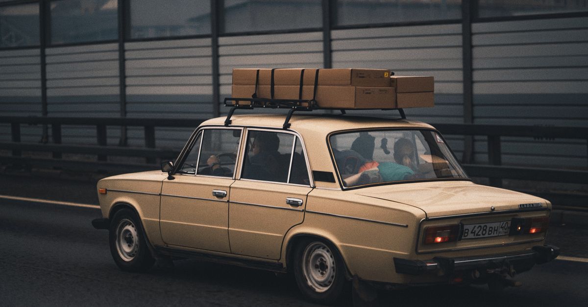 Classic beige sedan carrying luggage, driving on highway with city in background.