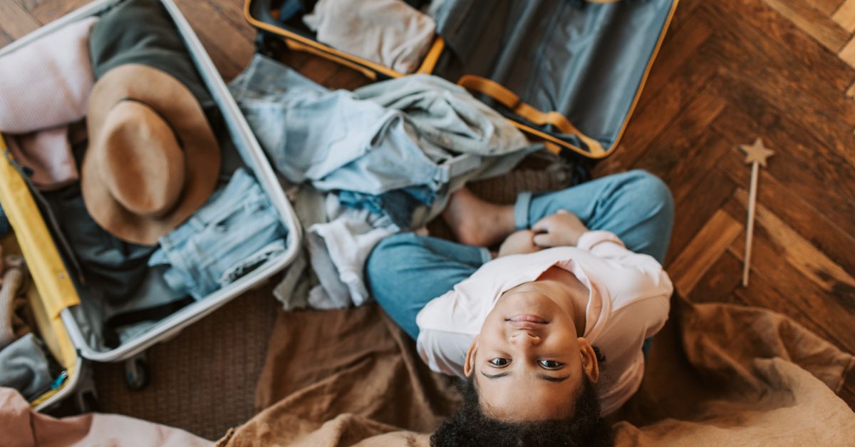 Young child surrounded by clothes and an open suitcase, preparing for a trip indoors.
